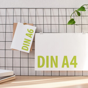 Zwei Drucksachen in den DIN-Formaten A4 und A6 hängen an Wandgitter, davor steht eine Tischlampe.