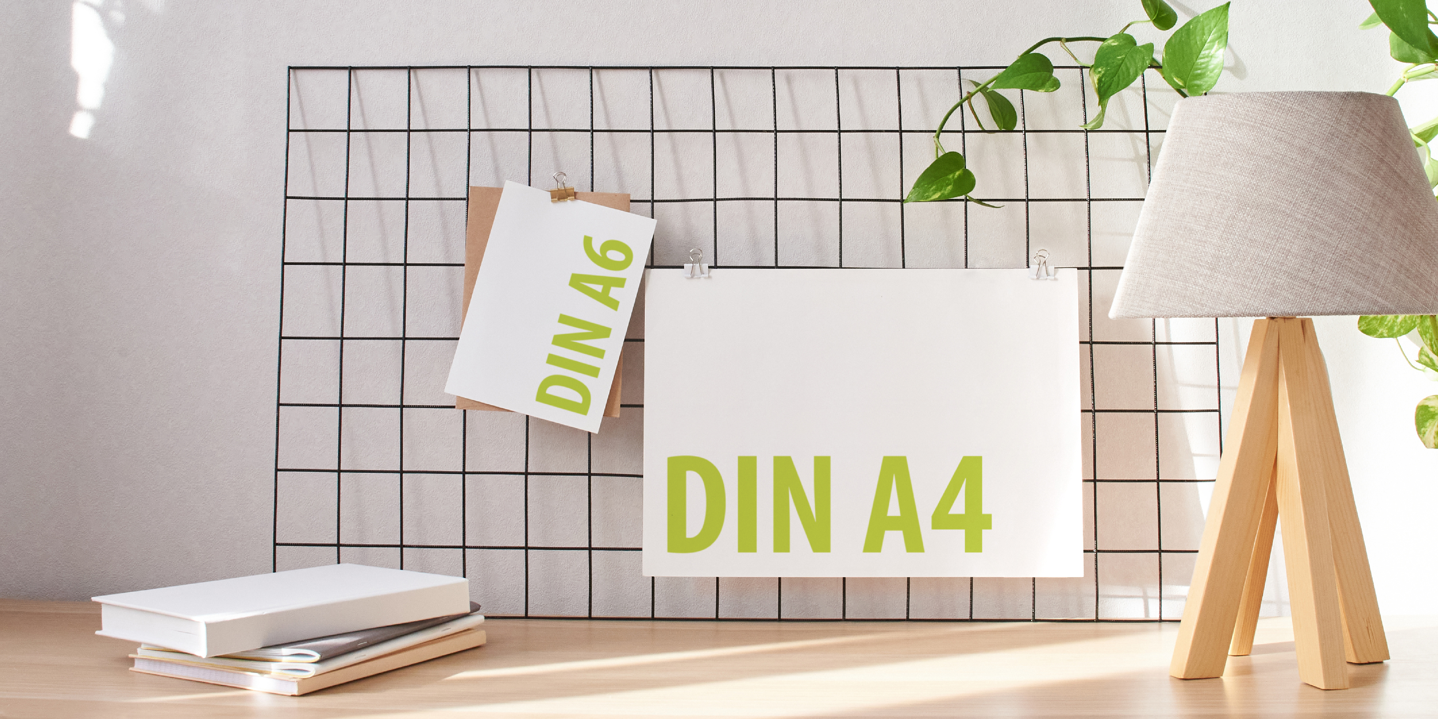 Zwei Drucksachen in den DIN-Formaten A4 und A6 hängen an Wandgitter, davor steht eine Tischlampe.
