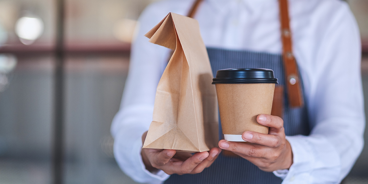 Verpackungsarten: Person in Café mit Schürze reicht Tüte mit Essen und Coffee-to-go-Becher