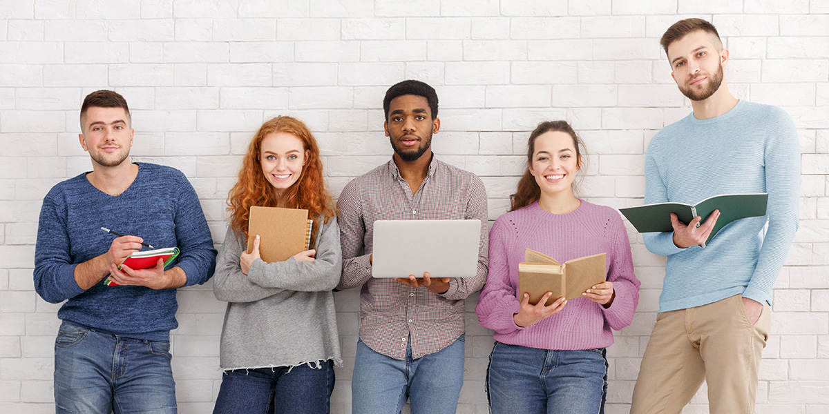 Fünf Personen lächeln, gucken in die Kamera & halten Bücher, Hefte & einen Laptop in Händen