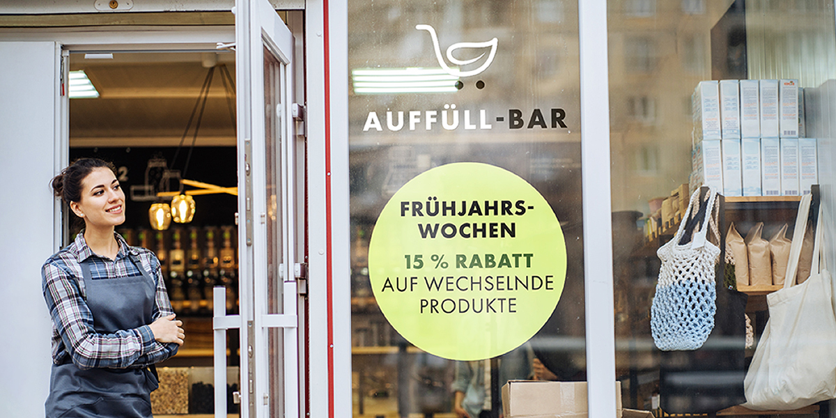 Lächelnde Person vor Schaufensterbeklebung mit Logo und Angebotshinweis eines Unverpacktladens