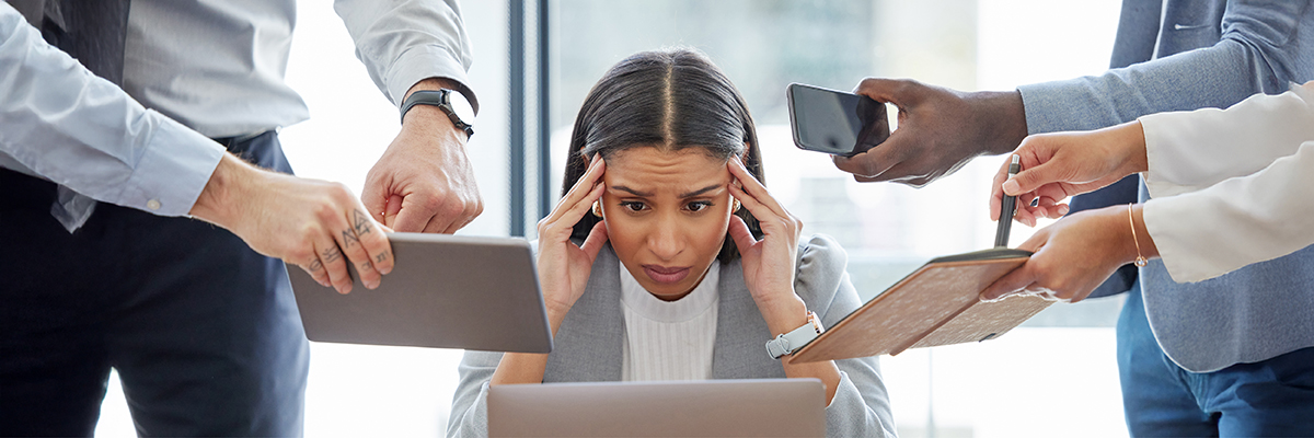 Gestresst aussehende Person sitzt am Laptop, drumherum stehen Leute mit Tablet, Handy & Notizbuch