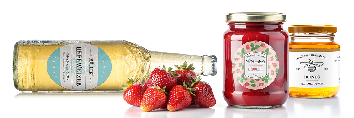 Bierflasche, Marmeladenglas und Honigglas, jeweils mit Etiketten; davor liegen frische Erdbeeren