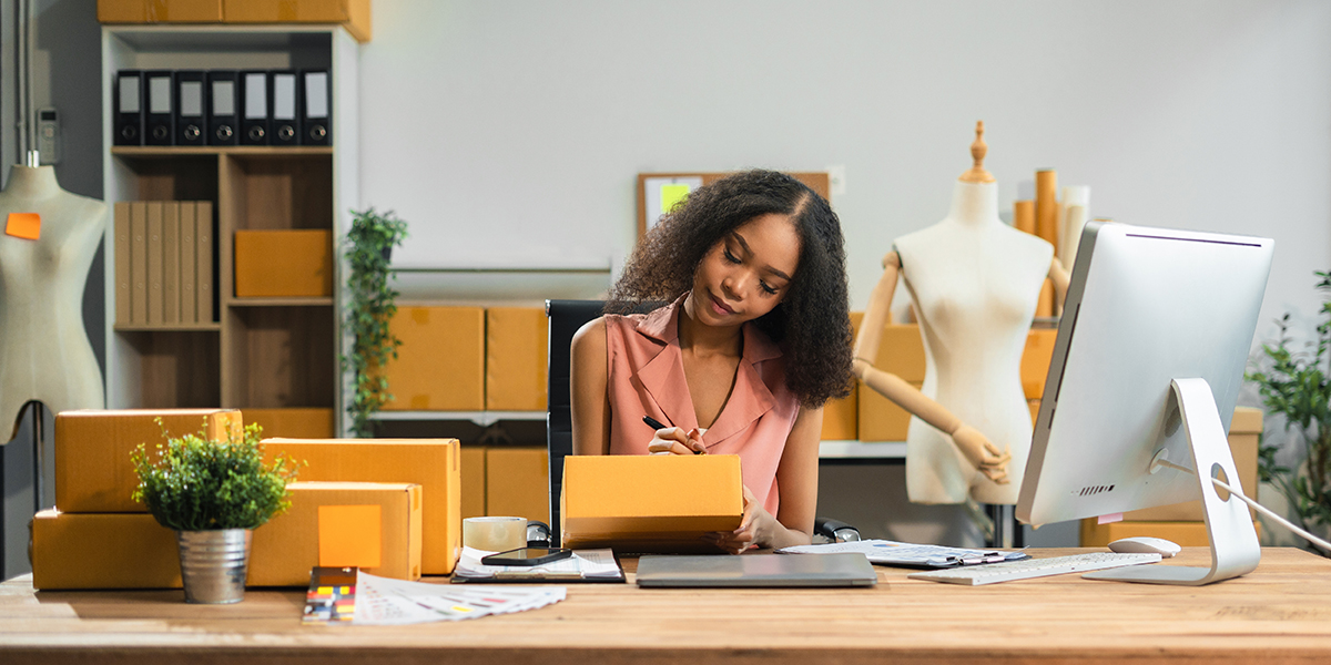 Eine Person sitzt am Schreibtisch und beschriftet Pakete, dahinter stehen Schneiderpuppen
