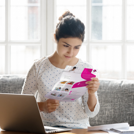 Eine Frau sitzt auf einem Sofa vorm Laptop und guckt ein Post-Mailing im pinkfarbenen Design an
