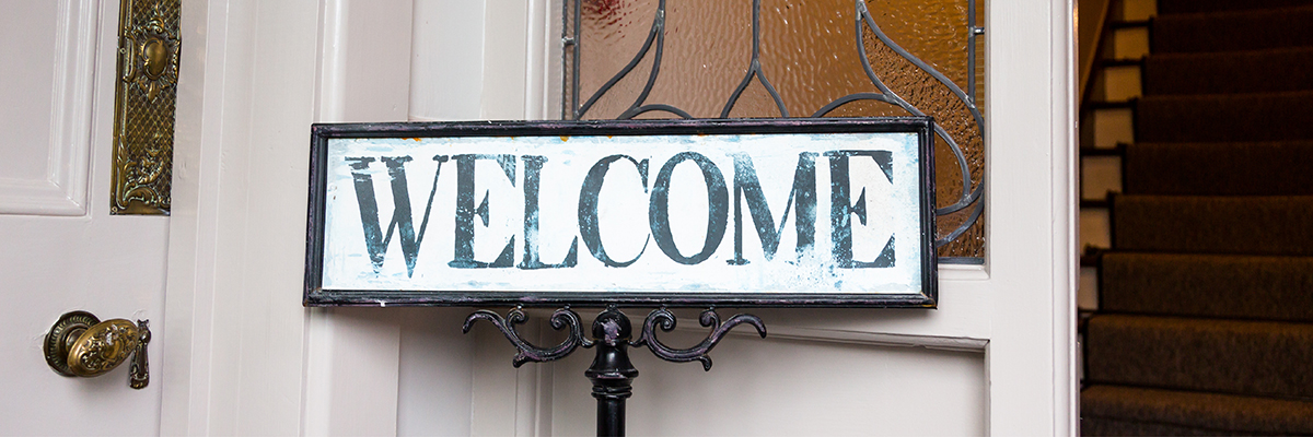 Vor einer weißen, schicken Haustür steht ein Schild mit der Aufschrift "WELCOME" in Serifenschrift