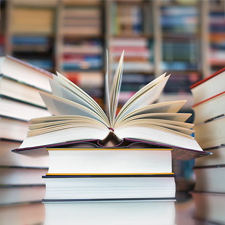 Ein Bücherstapel vor einem Bücherregal und weiteren Bucherstapeln, darauf ein aufgeschlagenes Buch