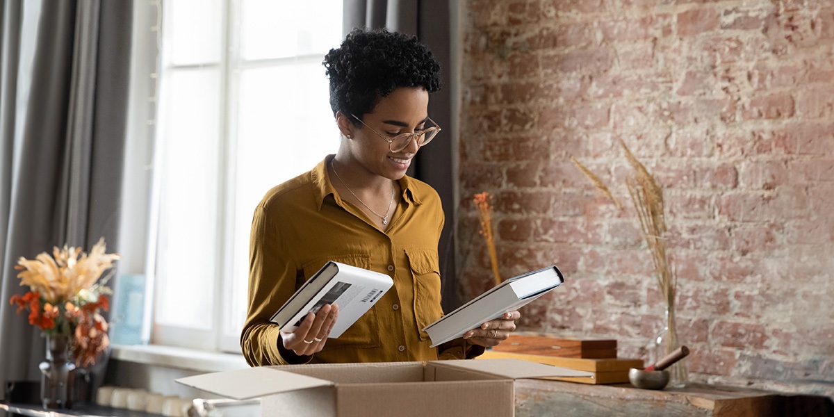 Lächelnde Autorin im Selfpublishing, zwei ihrer Bücher haltend über einem Karton