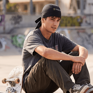 Ein Mann sitzt neben einem Skateboard und trägt individuell bedruckte Bandanamütze auf dem Kopf